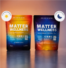 Two stand-up pouches of Matter Wellness supplement blends on a reflective surface against a warm gradient background. The left pouch is labeled Sunrise Blend Blueberry Flavour and has a red-to-blue gradient.