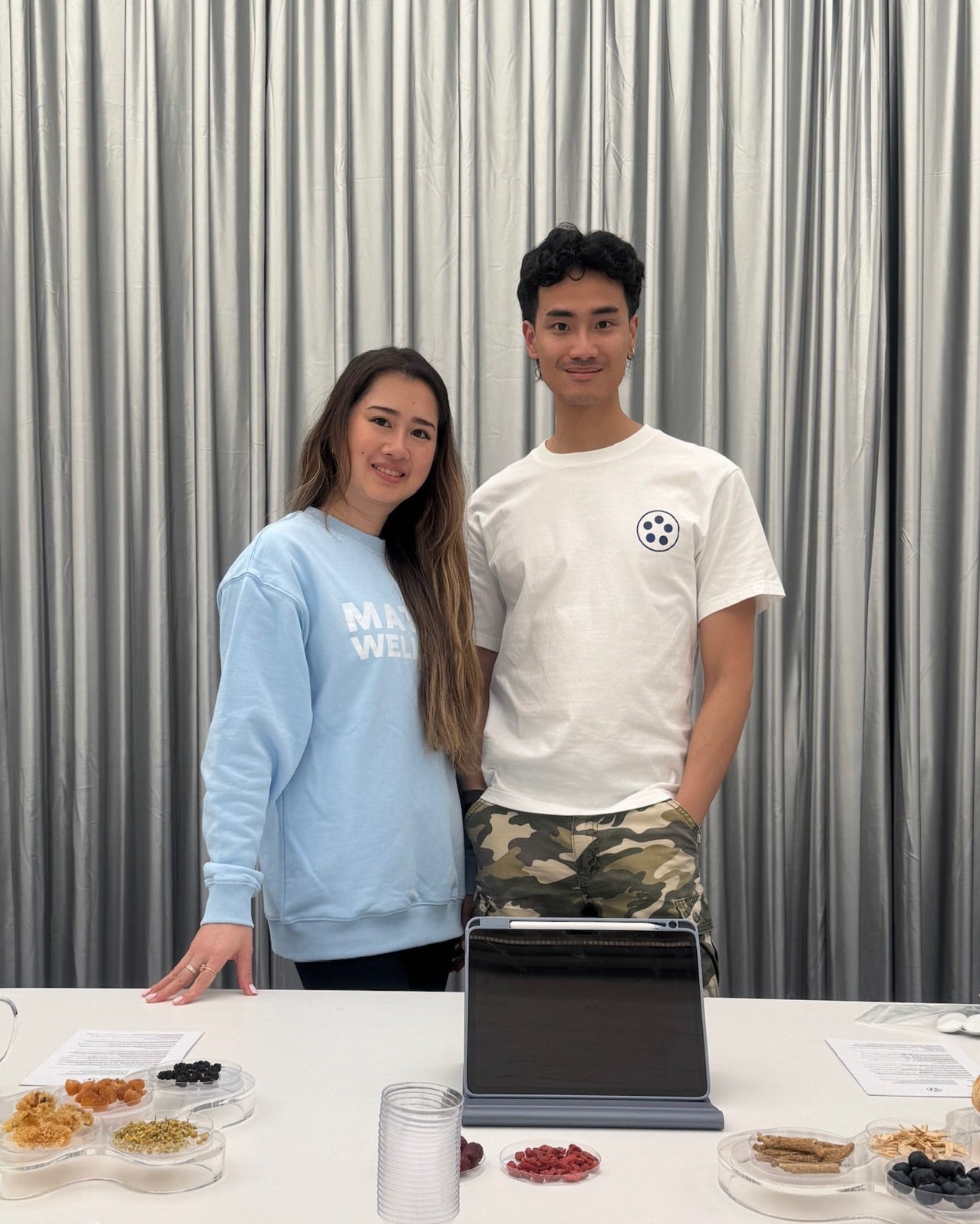Two founder standing behind a table with food and a laptop against a silver curtain background
